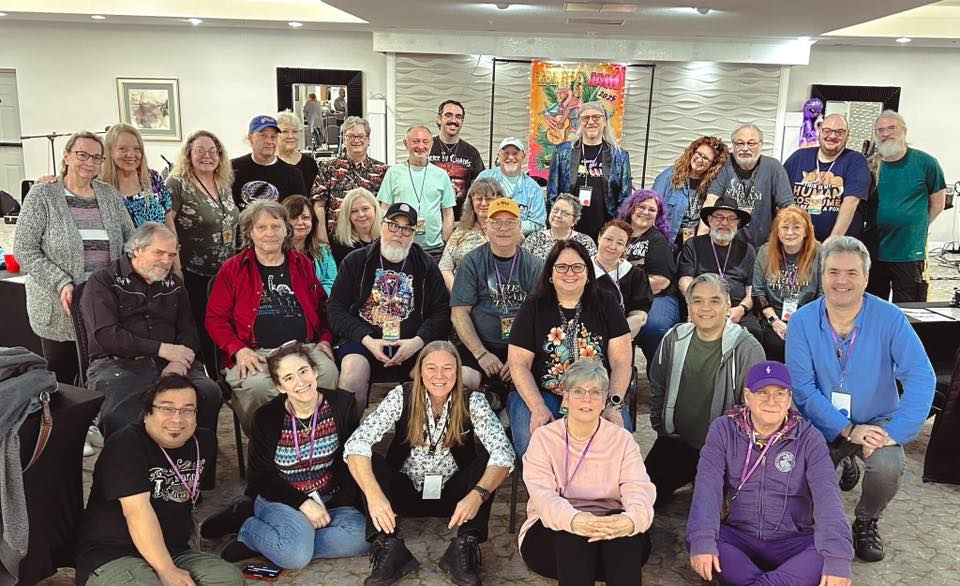 Group of diverse SL Live Music Jams performers gathered for a photo at an indoor event, with some wearing name tags and lanyards, and a colorful backdrop in the background.